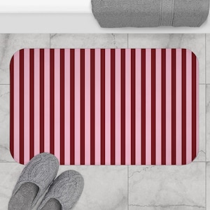 Puede incluir: Una alfombra de baño rectangular con un patrón de rayas verticales en tonos burdeos y rosa claro alternados. La alfombra está colocada sobre un suelo embaldosado, con un par de zapatillas de punto grises en la parte inferior. Una toalla gris es visible en el fondo.