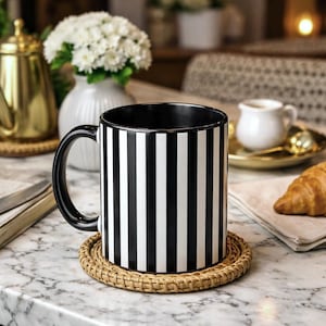 Pode incluir: Uma caneca de cerâmica listrada em preto e branco com uma alça preta está em uma base redonda tecida. A caneca está cheia de um líquido escuro. Uma chaleira dourada e um vaso branco com flores estão ao fundo.