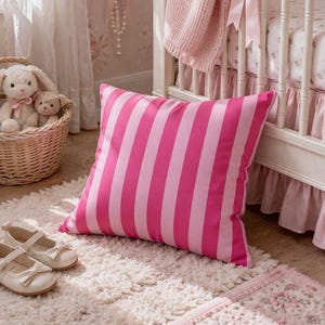 May include: A cushion with pink and white stripes sits on a fluffy white rug in a nursery. The room includes a cot with a pink blanket and a basket with soft toys. A pair of white shoes with bows are also visible.
