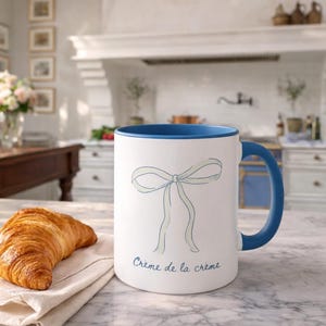 May include: White ceramic mug with a blue interior and handle. The mug features a light blue bow design and the text "Crème de la crème". A croissant rests on a white napkin on a marble countertop.