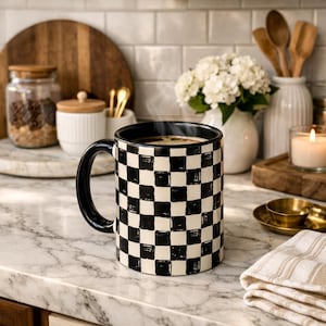 Puede incluir: Una taza de café a cuadros blancos y negros con un asa negra, llena de una bebida caliente. La taza está sobre una encimera de mármol, con una toalla a rayas de tonos neutros y otros artículos de cocina en el fondo.