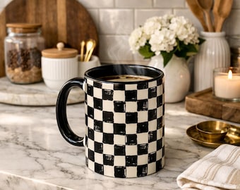 May include: A black and white checkered coffee mug with a black handle, filled with a hot beverage. The mug sits on a marble countertop, with a neutral-toned striped towel and other kitchen items in the background.