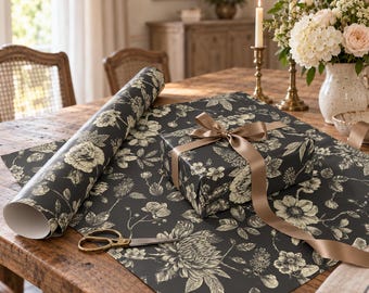 Puede incluir: Un rollo de papel de regalo gris oscuro con un estampado floral botánico en color crema, una caja de regalo rectangular envuelta con el mismo papel y un lazo de satén, junto con unas tijeras de metal de tono dorado.