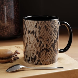 May include: A ceramic coffee mug with a snakeskin pattern in shades of brown and tan. The interior and handle are black. A silver spoon, a macaron, and coffee beans are on a wooden surface.