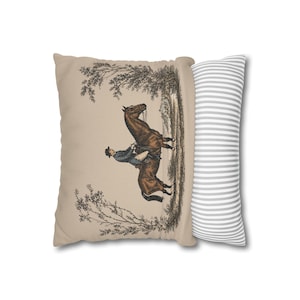 May include: A rectangular throw pillow featuring a beige front panel with a detailed illustration of a person riding a brown horse through a wooded landscape. The side panel of the pillow displays a pattern of thin, alternating white and gray stripes.