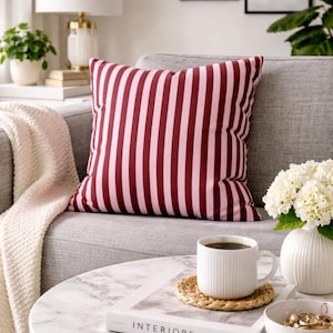 Op de afbeelding: Een decoratief kussen met verticale roze en bordeauxrode strepen op een grijze bank. Een witte koffiemok staat op een marmeren tafel met een boek met de tekst "INTERIORS". Een witte vaas bevat witte bloemen.