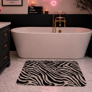 May include: A rectangular zebra-print bath mat in black and beige sits on a marble floor in front of a white bathtub. The bathroom has a black accent wall, gold fixtures, and a neon heart sign.