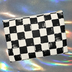 Peut inclure: Une trousse de maquillage rectangulaire avec un motif de damier noir et blanc. Le sac a une fermeture éclair dorée et un fond crème. Les carrés noirs ont un aspect texturé au pinceau.