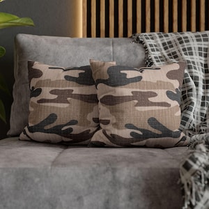 May include: Two square throw pillows with a camouflage pattern in shades of brown, tan, and black. The pillows are resting on a gray sofa. A plaid blanket is draped over the back of the sofa.