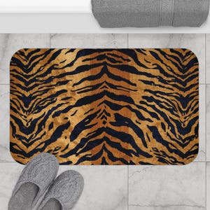 Op de afbeelding: Een rechthoekige badmat met een tijgerstreepmotief in bruin en zwart. De mat ligt op een betegelde vloer, met een paar grijze pantoffels en een grijze handdoek zichtbaar. Het tapijt heeft afgeronde hoeken.