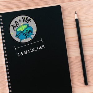 May include: A black notebook with a spiral binding and a round sticker on the front. The sticker is gray with a green cauldron, stars, and the text "Turn to Page 394". The notebook is 2 and 3/4 inches wide.