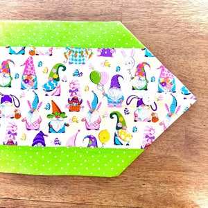 Może przedstawiać: Zielony i biały bieżnik stołowy z wzorem krasnoludków i jajek wielkanocnych. Bieżnik ma zieloną obwódkę w kropki.