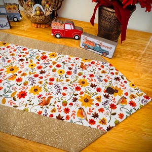 May include: A fall-themed table runner with a white background, featuring sunflowers, leaves, squirrels, and birds. The runner has a tan border with a swirl pattern and a wooden button. Decorative fall figurines and flowers are in the background.