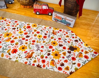 May include: A fall-themed table runner with a white background, featuring sunflowers, leaves, squirrels, and birds. The runner has a tan border with a swirl pattern and a wooden button. Decorative fall figurines and flowers are in the background.