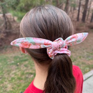 Pode incluir: Um scrunchie para cabelo com padrão floral rosa e verde com um toque de brilho dourado. O scrunchie está amarrado em torno de um rabo de cavalo.