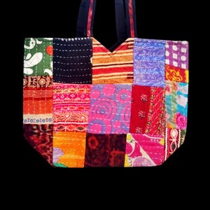 Puede incluir: Un bolso tote de patchwork con una variedad de cuadrados de tela coloridos. El bolso presenta una mezcla de patrones, incluyendo rayas, diseños florales y formas abstractas. Las asas son de color azul oscuro y el bolso está sobre un fondo negro.