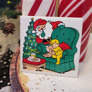 May include: A square ceramic tile with a Christmas scene featuring Santa Claus, a child, and a dog. The tile is set in front of two red and white striped candles. A small table has a glass of milk and cookies. A Christmas tree is in the background.