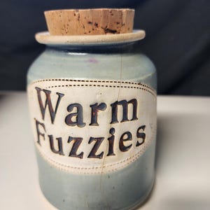 May include: A small, light blue ceramic jar with a cork stopper. The jar has a white label with the words "Warm Fuzzies" in brown lettering. The jar has a crack running down the side.