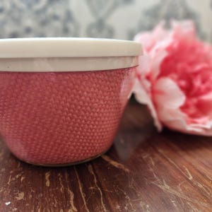May include: A small, pink container with a textured, woven-like exterior and a white lid. A large, pink peony flower is blurred in the background. The container sits on a wooden surface.