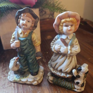 May include: Two porcelain figurines depicting children. The left figure wears a dark hat, yellow shirt, and green overalls, standing with a white duck. The right figure wears a pink hat and white dress, standing with a small black and white dog.