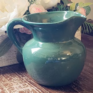 May include: A solid dark green ceramic pitcher with a rounded body, a wide flared rim, and a curved handle on the side. The surface of the pitcher has a smooth, glossy finish that reflects light, creating bright highlights across the curved exterior.