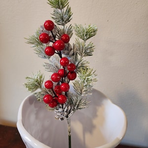 May include: A sprig of artificial greenery with red berries, displayed in a white, decorative vase. The faux pine needles are dusted with a snowy effect, and the bright red berries provide a festive contrast. The vase has a unique, textured design.
