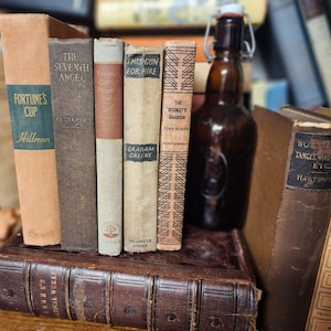 May include: A collection of vintage books with aged covers, including titles like "Fortune's Cup" and "The Seventh Angel." A brown glass bottle with a swing top sits among the books. The books are stacked on a larger, leather-bound volume.