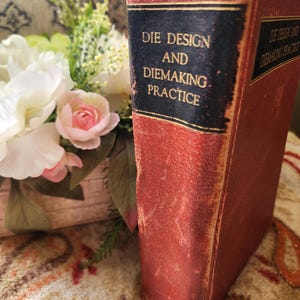 May include: A vintage book titled "Die Design and Diemaking Practice" with a worn, red cover and a black label. The book is positioned next to a bouquet of white and pink flowers, suggesting a classic or antique theme.