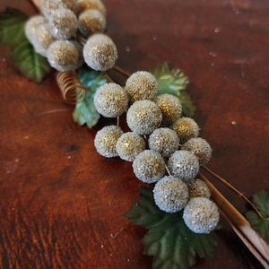 May include: A decorative floral stem featuring clusters of round, glitter-covered spheres in shades of gold and silver. Green fabric leaves and a brown stem complete the design, set against a dark wooden surface.