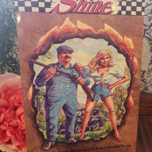May include: A book cover with the title "Shine" in a stylized font. The cover art depicts a man in overalls holding a rifle and a woman in denim shorts. The background is a brown, textured design with a fiery border. The text reads "A Story of Moonshine, Murder and the Birth of Southern Racing by Rodger Doss."
