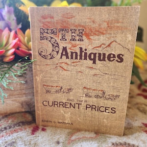 May include: A vintage book titled "5th Antiques and Their Current Prices" with a tan cover. The cover features illustrations of covered wagons and mountains in brown and orange hues. The author's name, Edwin G. Warman, is printed at the bottom.