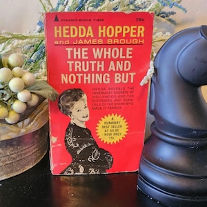 May include: A vintage paperback book titled "HEDDA HOPPER and JAMES BROUGH: THE WHOLE TRUTH AND NOTHING BUT." The cover is red with black and yellow text, featuring a portrait of a woman. A black chess piece is on the right.