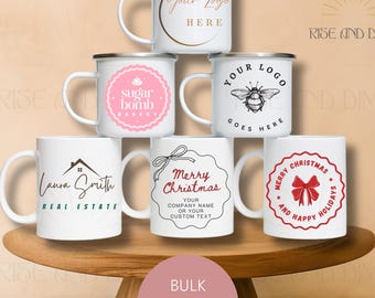May include: A collection of six white ceramic and enamel-style mugs featuring various custom designs, including logos, text, and holiday graphics. The mugs are arranged in a pyramid stack, displaying prints like 'sugar bomb BAKERY', 'Laura Smith REAL ESTATE', and 'Merry Christmas'.