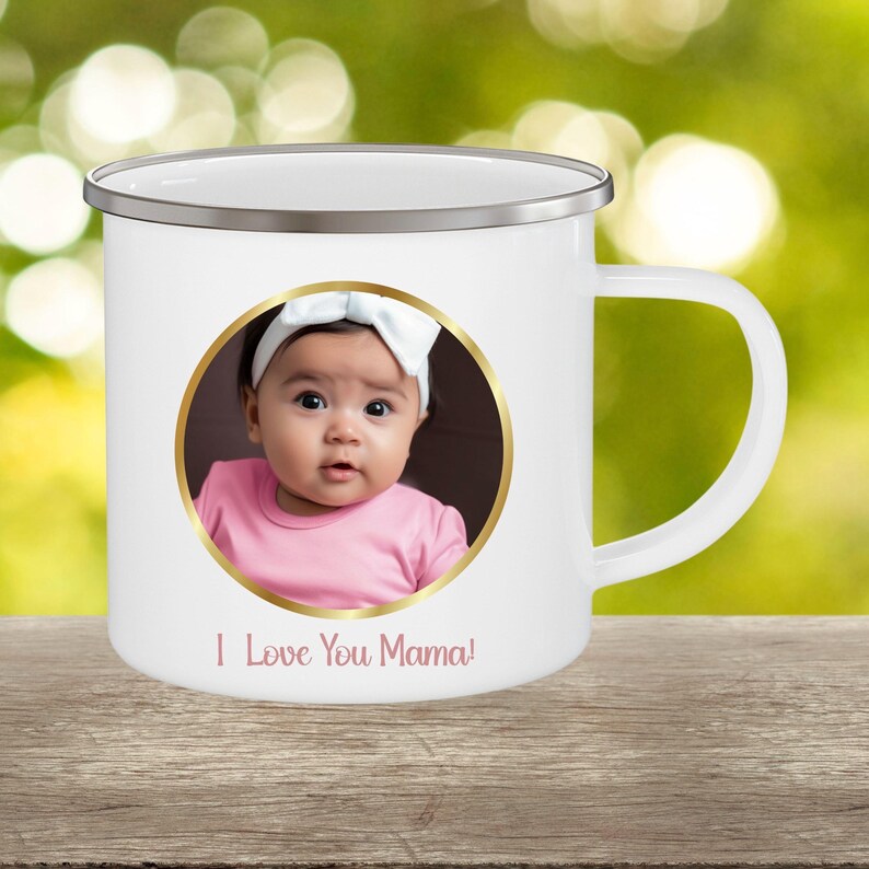 May include: A white enamel mug with a silver rim and a handle on the right side. The front features a circular gold-framed photograph of a baby wearing a white headband and a pink shirt. Below the photo, the text reads I Love You Mama! in pink.