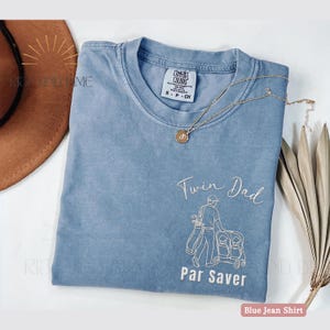 May include: A blue jean colored t-shirt with the words "Twin Dad" and "Par Saver" in white script. The shirt features a graphic of a golfer pushing a double stroller. A gold necklace with a pendant rests on the shirt.