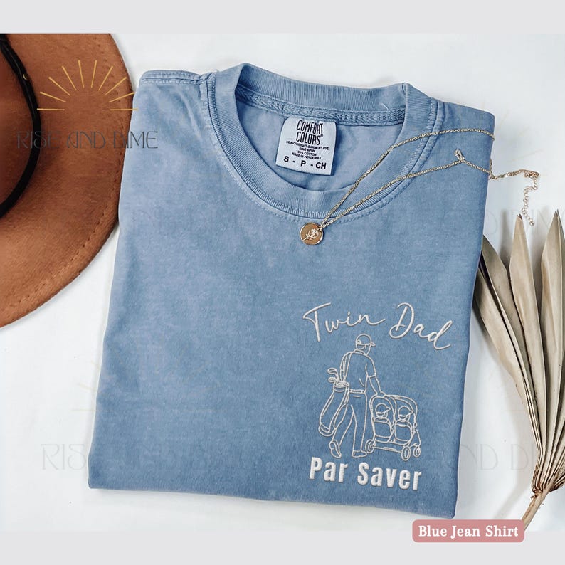May include: A blue jean colored t-shirt with the words "Twin Dad" and "Par Saver" in white script. The shirt features a graphic of a golfer pushing a double stroller. A gold necklace with a pendant rests on the shirt.