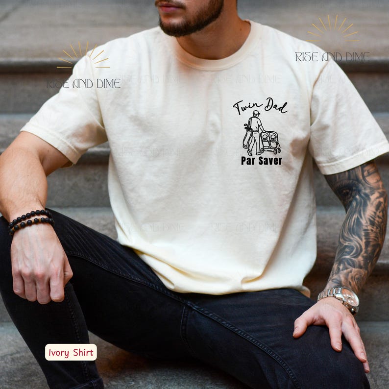 May include: Ivory-colored t-shirt with the text "Twin Dad Par Saver" and a graphic of a golfer with a golf cart. The shirt is a short-sleeved crew neck. The model is wearing black jeans and a watch.