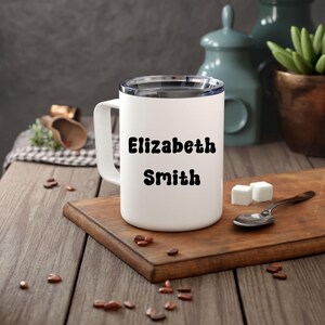 May include: A white cylindrical mug with a handle and a clear lid, featuring the black text Elizabeth Smith printed in a bold, rounded font on the front surface. The mug is constructed from a smooth material and has a simple, functional design.