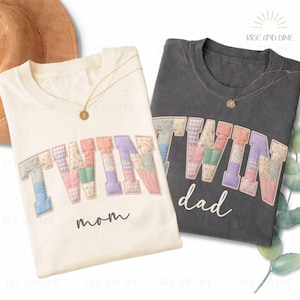 May include: Two t-shirts, one cream and one charcoal, with the word "TWIN" in patchwork letters. The cream shirt says "mom" below the word, and the charcoal shirt says "dad". Both shirts have gold necklaces.