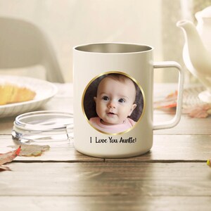 May include: A cream-colored mug with a handle and a circular gold-rimmed frame containing a photograph of a baby. Below the circular frame, the text "I Love You Auntie!" is printed in a black script font. The mug has a smooth, matte finish.