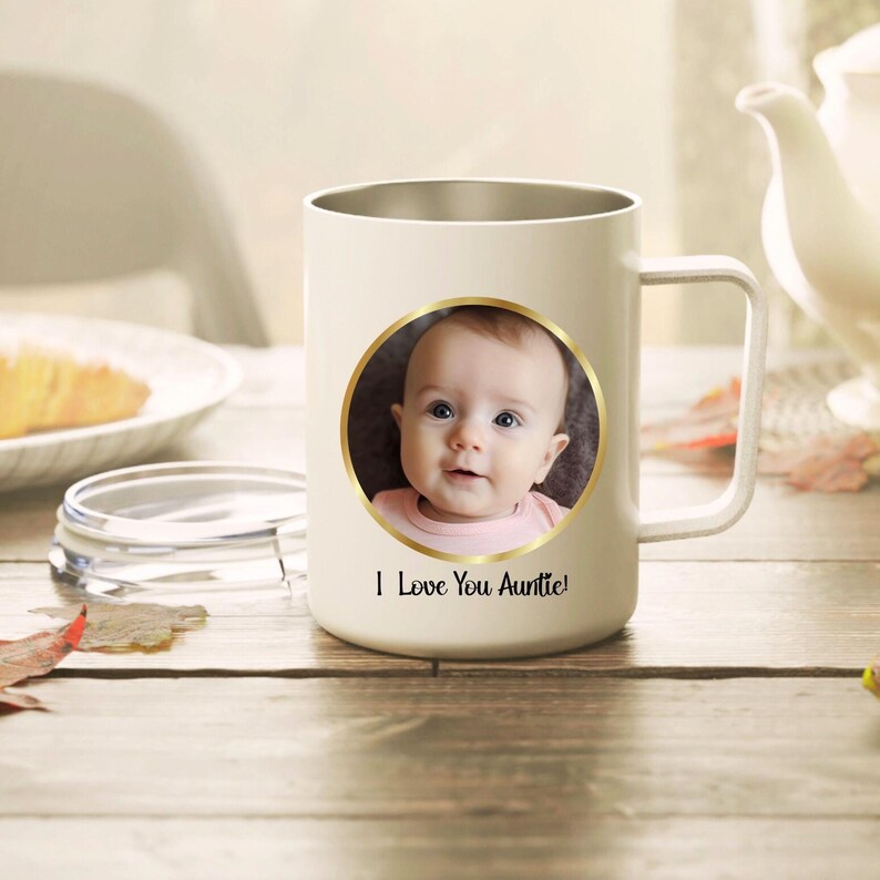 May include: A cream-colored mug with a handle and a circular gold-rimmed frame containing a photograph of a baby. Below the circular frame, the text "I Love You Auntie!" is printed in a black script font. The mug has a smooth, matte finish.