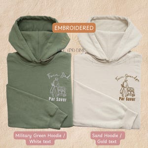May include: Two hooded sweatshirts, one military green and one sand-colored. Both feature embroidered text and a golf-themed graphic. The military green hoodie has white text that reads "Twin Dad" and "Par Saver." The sand hoodie has gold text that reads "Twin Grandpa" and "Par Saver."
