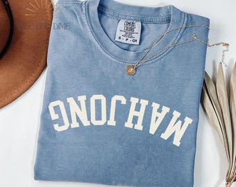 May include: A blue jean-coloured t-shirt with the word "MAHJONG" printed in white block letters. The shirt has a crew neck and short sleeves. A gold necklace with a pendant is draped across the shirt.