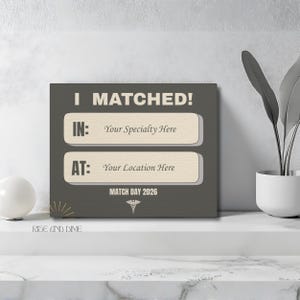 K&ouml;nnte beinhalten: Ein quadratischer Leinwanddruck mit dem wei&szlig;en Text "I MATCHED!". Darunter stehen in zwei rechteckigen Feldern "IN: Your Specialty Here" und "AT: Your Location Here". Der untere Text lautet "MATCH DAY 2026". Die Leinwand ist grau.