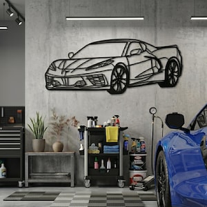 Puede incluir: Una pieza de arte mural de metal negro que representa un coche deportivo, montada en una pared de hormigón gris. El diseño del coche es detallado, con ruedas y contornos de la carrocería visibles. El garaje incluye un coche deportivo azul, un carro de herramientas y plantas.