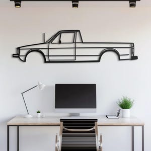 以下が含まれることがあります： 白い壁に、ピックアップトラックの黒い金属製のアウトラインが取り付けられています。その下には、コンピューター、ランプ、椅子のあるデスクがあります。