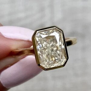 Pu&ograve; includere: Un anello d'oro con un grande diamante a taglio smeraldo, trasparente. Il diamante &egrave; incastonato in una montatura a bezel e ha una forma leggermente rettangolare.