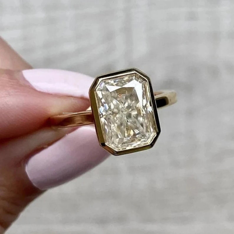 Pu&ograve; includere: Un anello d'oro con un grande diamante a taglio smeraldo, trasparente. Il diamante &egrave; incastonato in una montatura a bezel e ha una forma leggermente rettangolare.