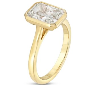 Pu&ograve; includere: Un anello d'oro con una grande pietra preziosa trasparente a taglio smeraldo. L'anello ha una fascia semplice e una montatura a  bezel.