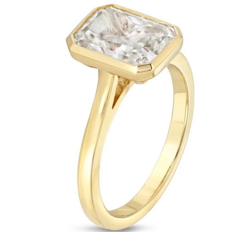 Pu&ograve; includere: Un anello d'oro con una grande pietra preziosa trasparente a taglio smeraldo. L'anello ha una fascia semplice e una montatura a  bezel.